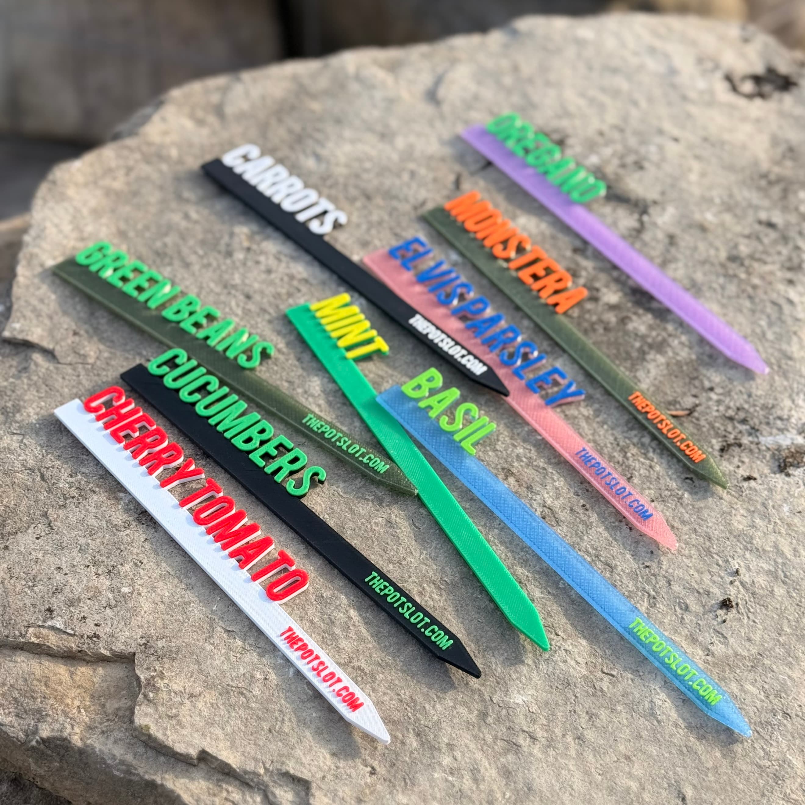 Collection of colorful 3D-printed garden picks scattered on a stone surface showing custom color combinations for Carrots, Mint, Green Beans, Basil, and Cherry Tomato