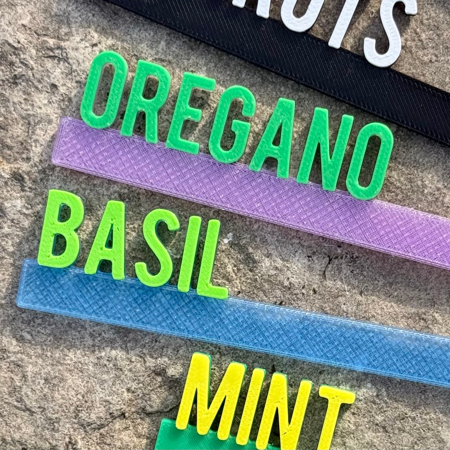 Close-up detail of raised 3D lettering on garden picks reading Oregano in green, Basil in yellow, and Mint in purple