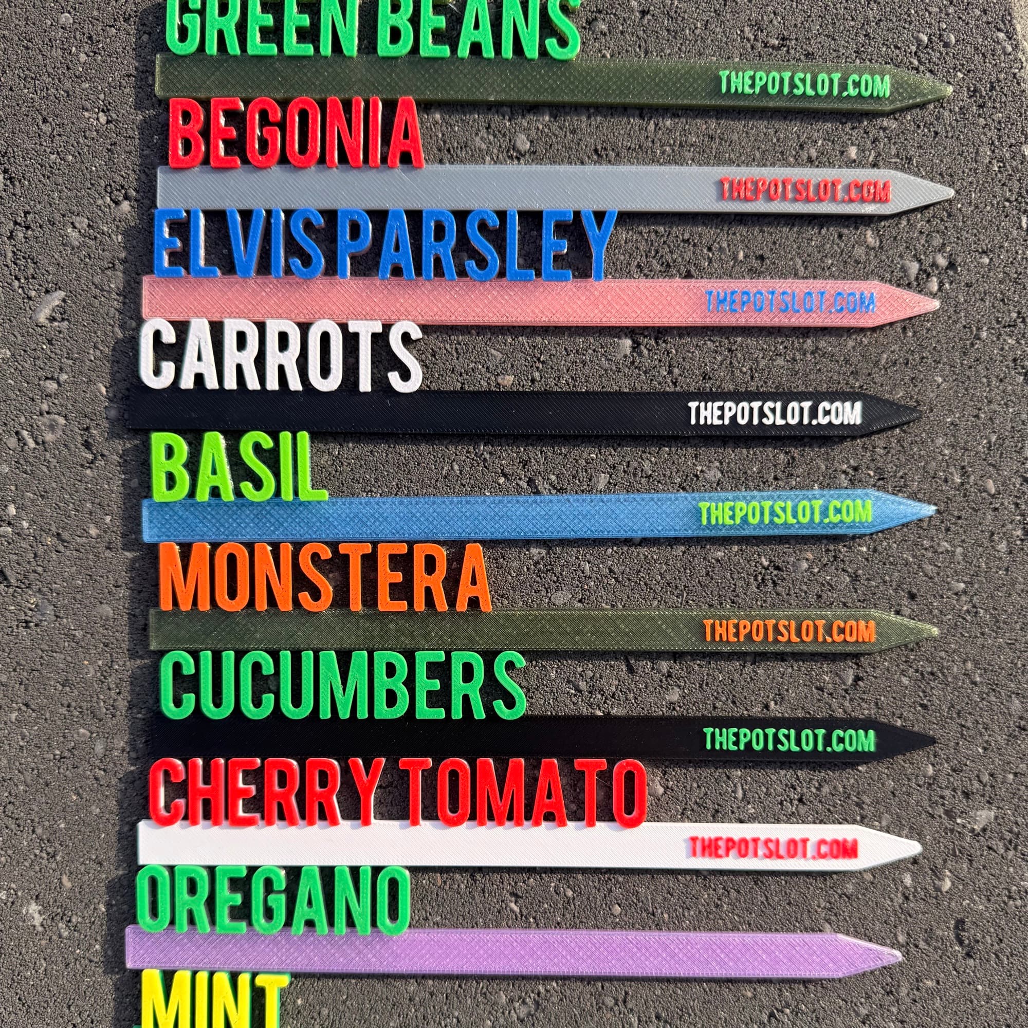 Row of 3D-printed garden picks in assorted colors with raised text showing Green Beans, Begonia, Elvis Parsley, Carrots, Basil, Monstera, Cucumbers, Cherry Tomato, Oregano, and Mint