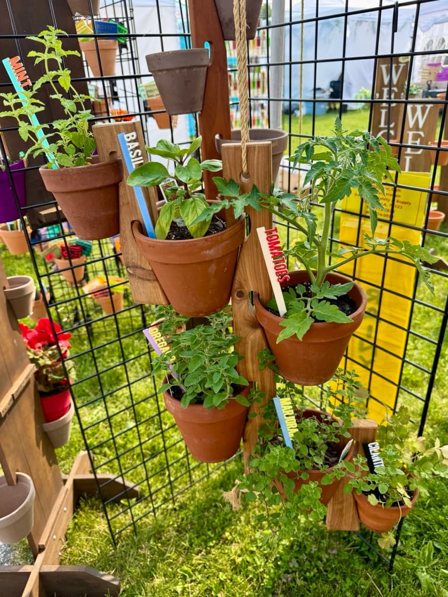 Wooden Pot Slot hanger with colorful 3D-printed garden picks labeled Basil, Tomatoes, and herbs in terra cotta pots at an outdoor farmers market