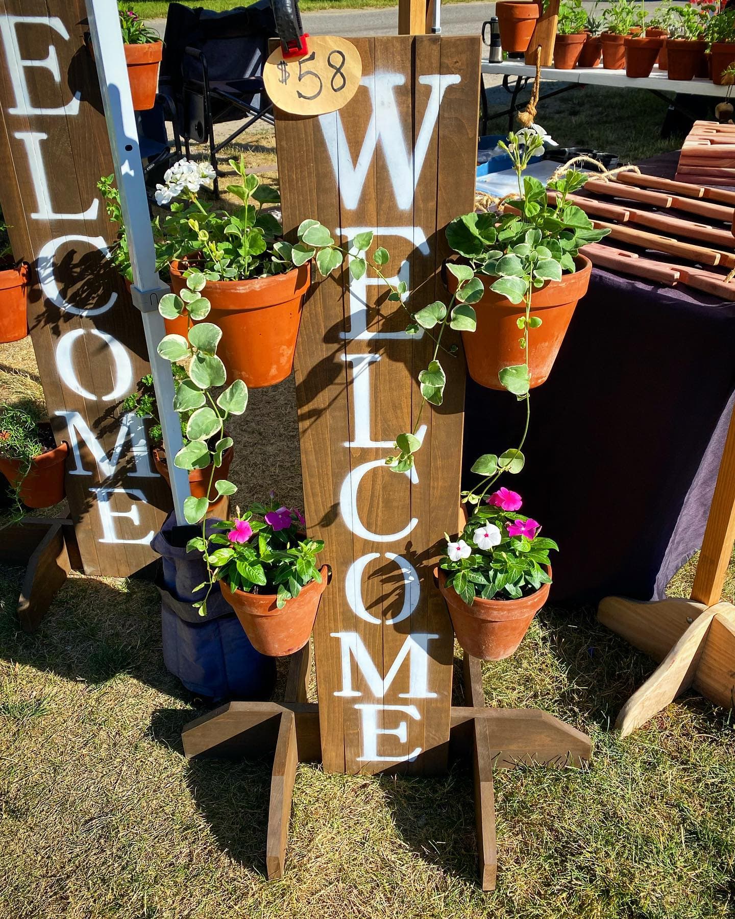 Handcrafted cedar welcome sign with routed letter cutouts holding terra cotta pots of flowers at a farmers market