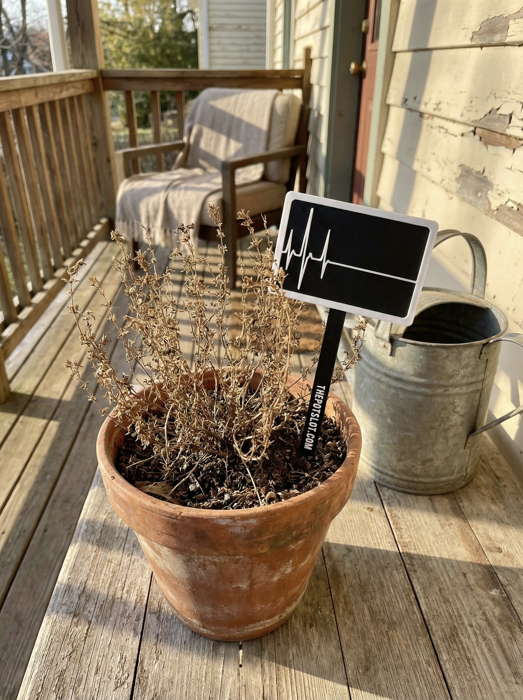 3D-printed pot pick in black and white with flatline heartbeat design in a terra cotta pot with a dead dried plant on a porch next to a watering can