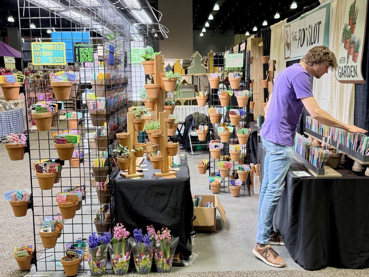 Will setting up The Pot Slot booth at an indoor craft show, organizing colorful 3D-printed garden picks and pot hangers